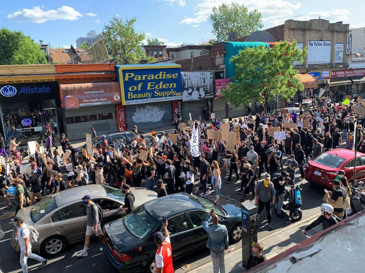 Image of a Black Lives Matter march. Crowd of people wearing masks and carrying signs walking down an urban street.