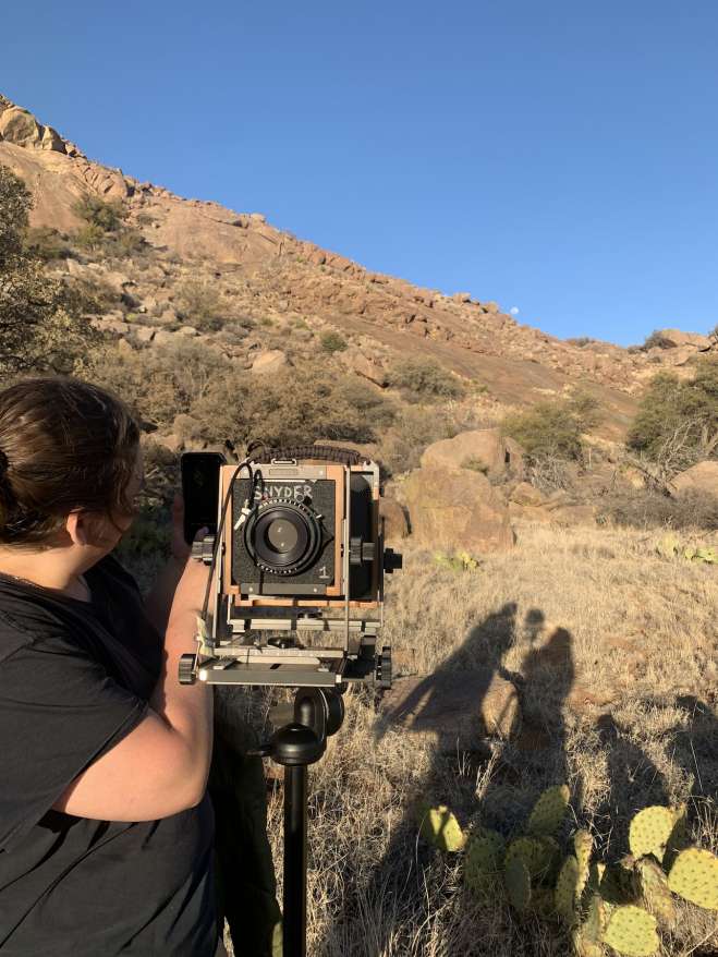 A person standing next to a large-formate camera in a desert landscape.