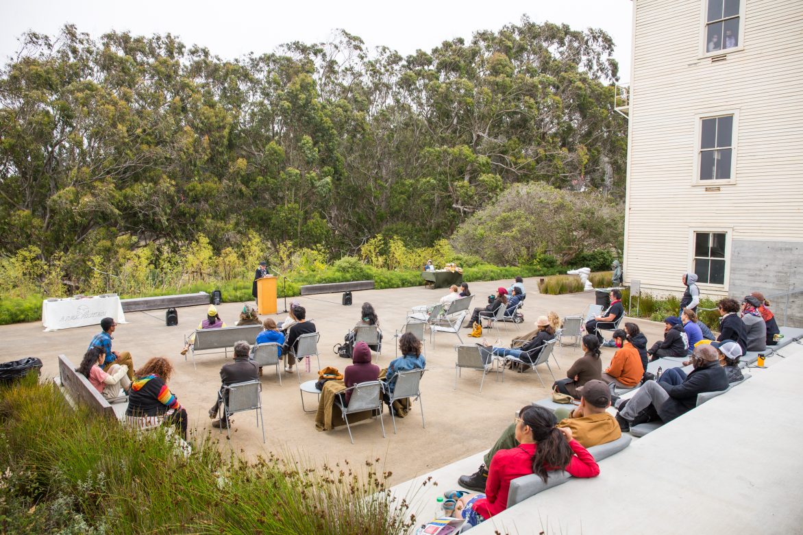 Image of an audience of people outdoors with a figure reading from a podium.