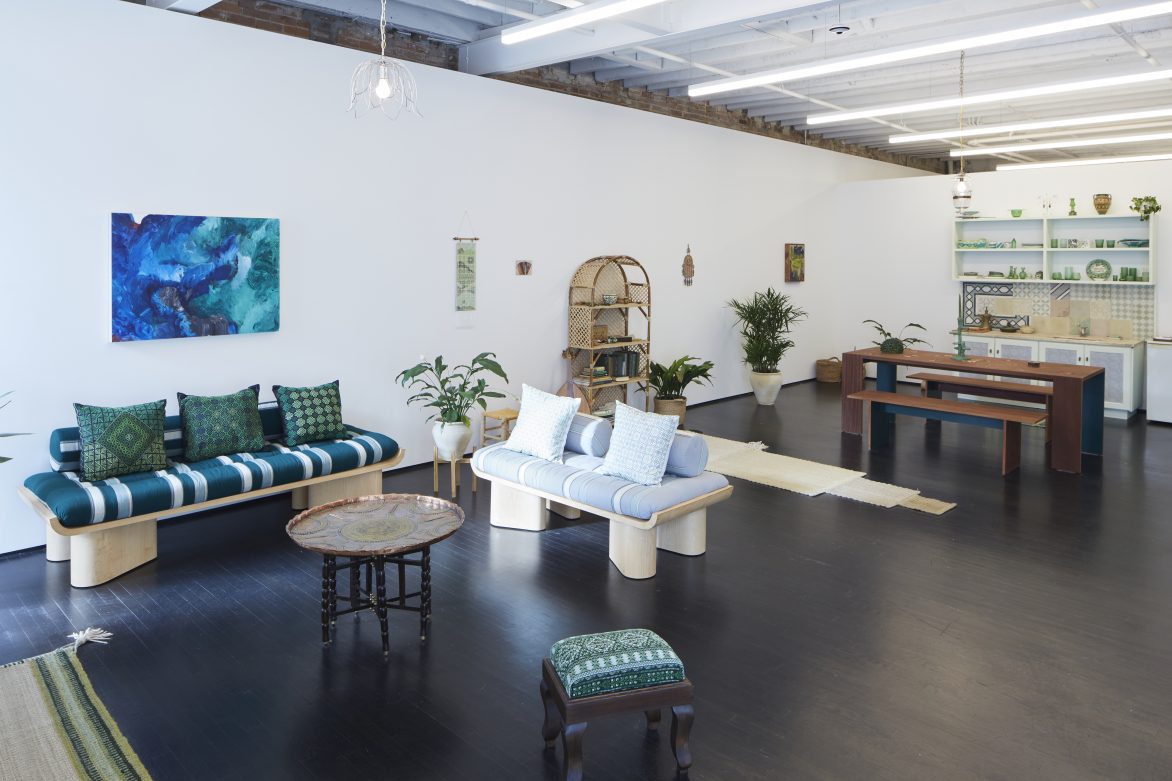 View of a gallery with an installation that looks like a domestic interior with sofas, coffee tables, plants, etc.