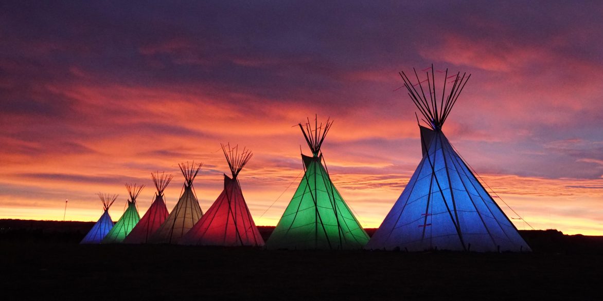 Image of teepees illuminate from inside with different colors against the sky at dusk.