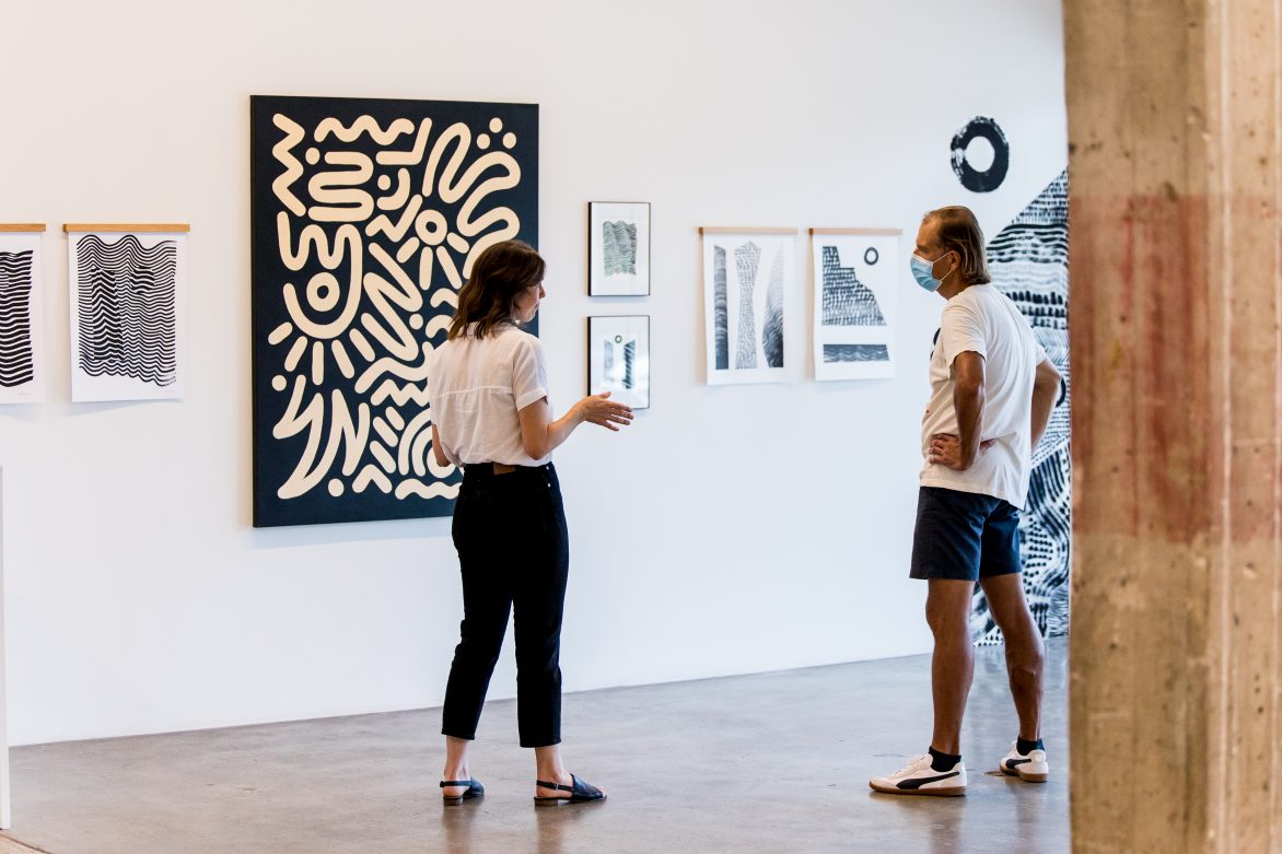 Two people in conversation standing in front of a wall with artworks.