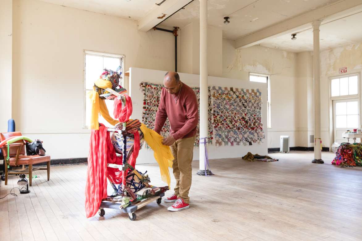 A man standing in a room, handling a piece of yellow fabric. Quilted fabric pieces are hung on a partition behind him and a heap of fabric is on the floor in the back left corner.