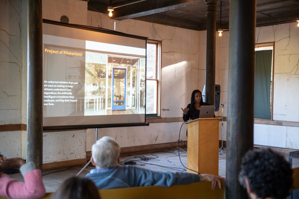 A woman standing at a podium address a group of people sitting in chairs. A project of an image with text is behind her.